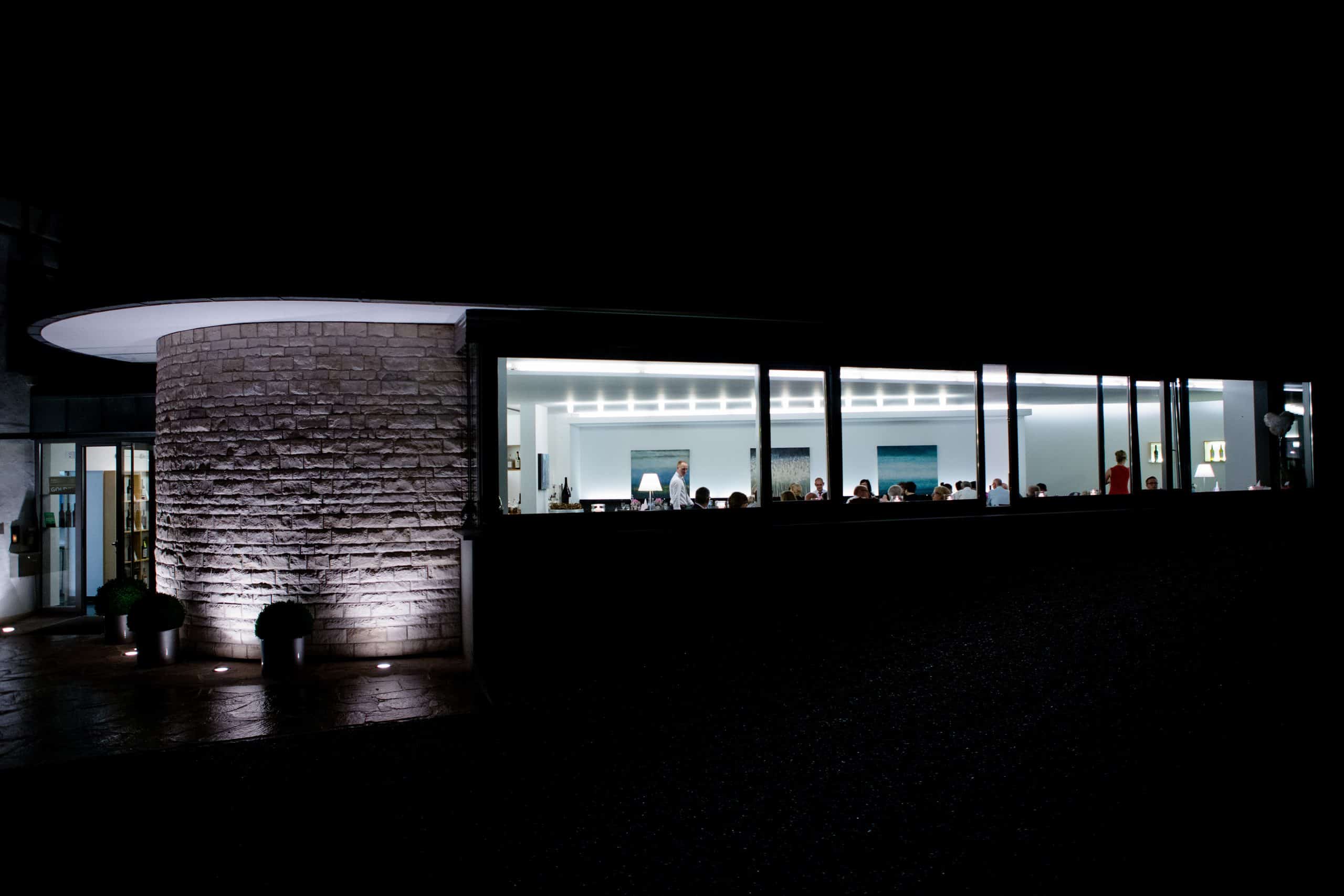 Blick von außen bei stockdunkler Nacht durch die hell beleuchteten Fenster des Weinforums. Innen sitzen viele Menschen an Tischen verteilt. Die Außenwand zum Eingang ist von unten beleuchtet.